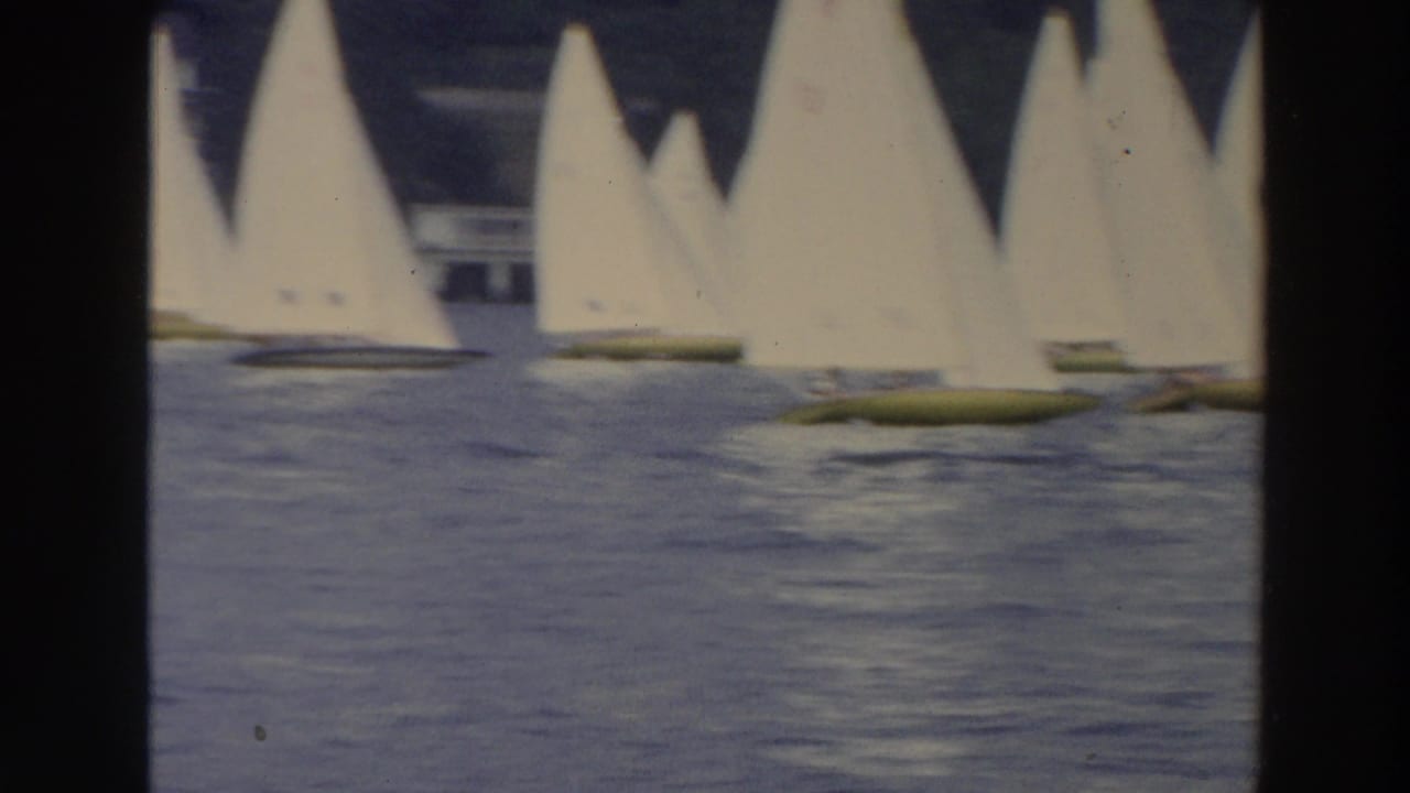 1966 Wisconsin home-movie frame with multiple white sails and yellow hulls grouped on calm water near a shaded shoreline.