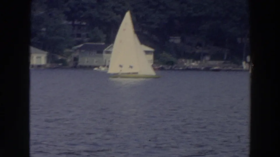 1966 Wisconsin home-movie frame of one small sailboat on calm water near a wooded residential shore.