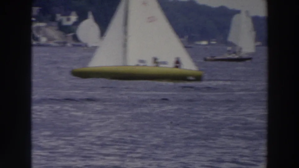 Closer 1966 Wisconsin lake view of a sailboat with a yellow hull and white sail, with other boats and hills behind it.