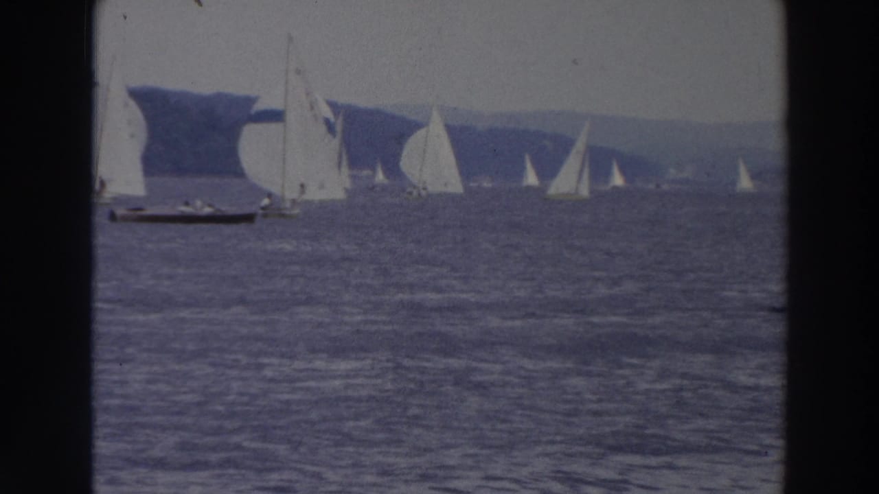 1966 Wisconsin home-movie frame with multiple sailboats and spinnakers racing on open bay water near a wooded shoreline.