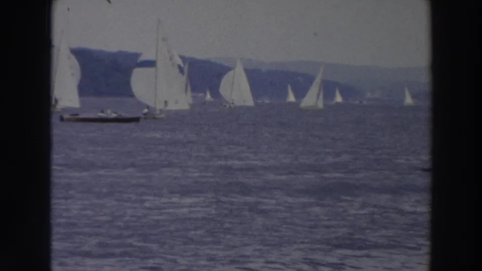 1966 Wisconsin home-movie frame with multiple sailboats and spinnakers racing on open bay water near a wooded shoreline.