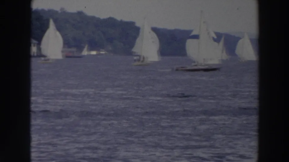 1966 Wisconsin 8mm frame showing a sailboat in the foreground with more racing sails along the tree-lined bay shore.