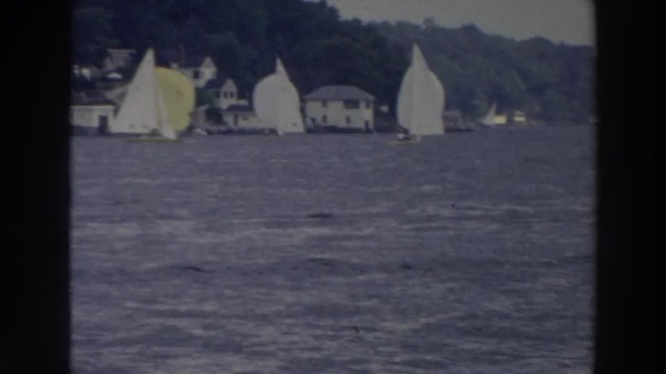 1966 Wisconsin 8mm frame of sailboats racing near waterfront buildings backed by a wooded hillside.