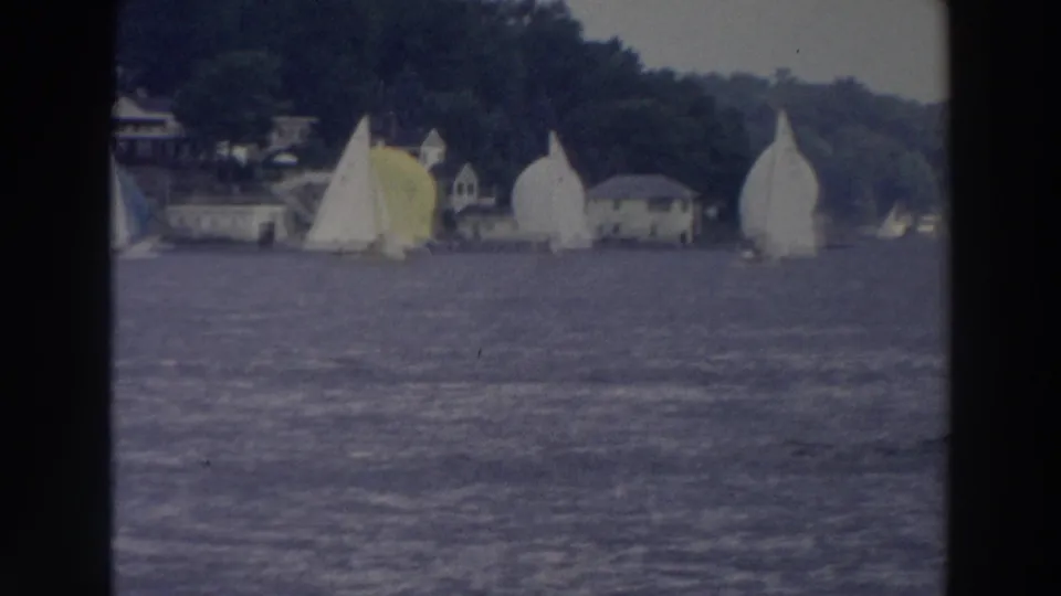 1966 Wisconsin 8mm frame showing small white sailboats crossing a blue lake near trees and low shoreline buildings.