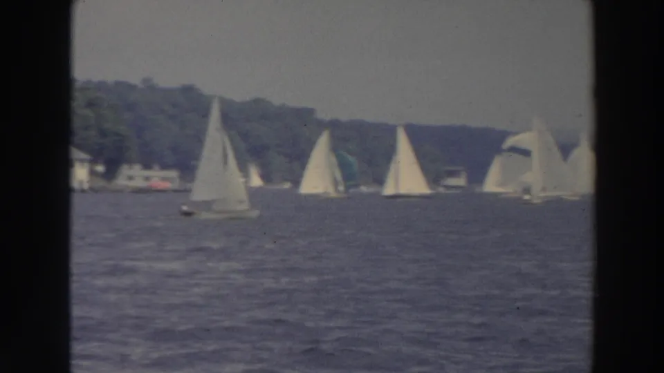 Wide 8mm frame of white sailboats dispersed across a Wisconsin lake, with a wooded shoreline beyond.