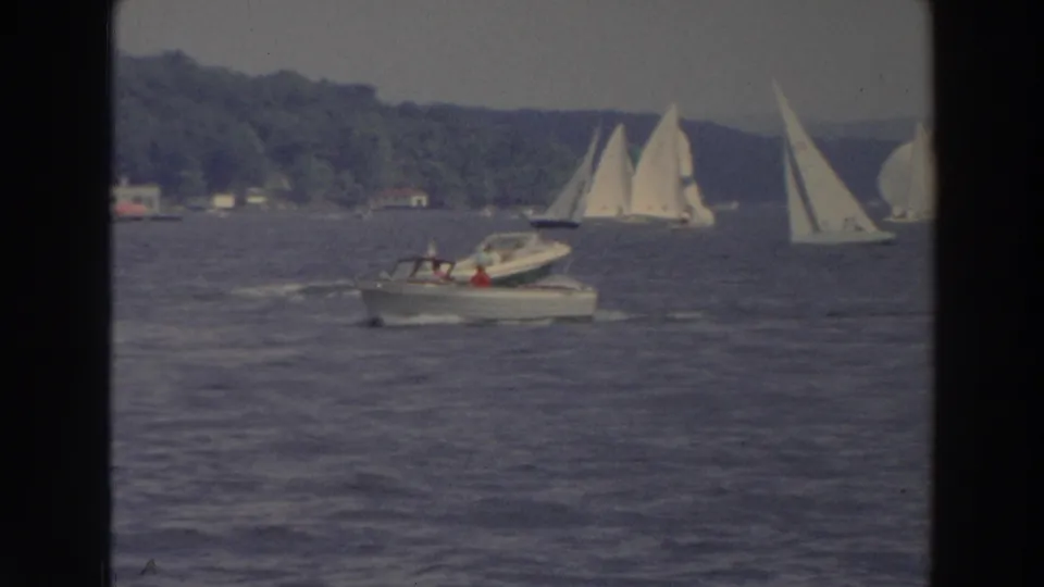 1966 Wisconsin 8mm frame with a small motorboat and nearby sailboats on a lake near a wooded shoreline.