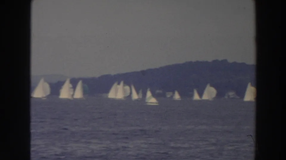 1966 Wisconsin 8mm still showing white sailboats stretched across lake water before a dark wooded hillside.