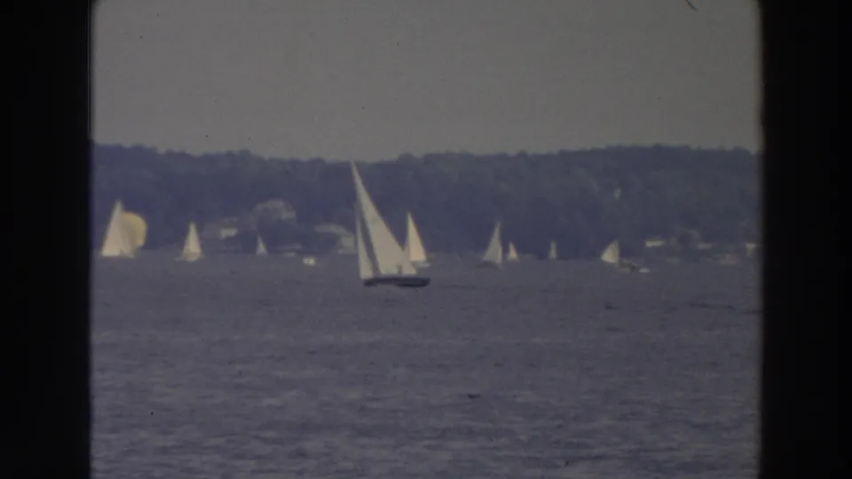 1966 Wisconsin lake footage with a nearer white-sailed boat in front of distant sailboats and a wooded far shore.