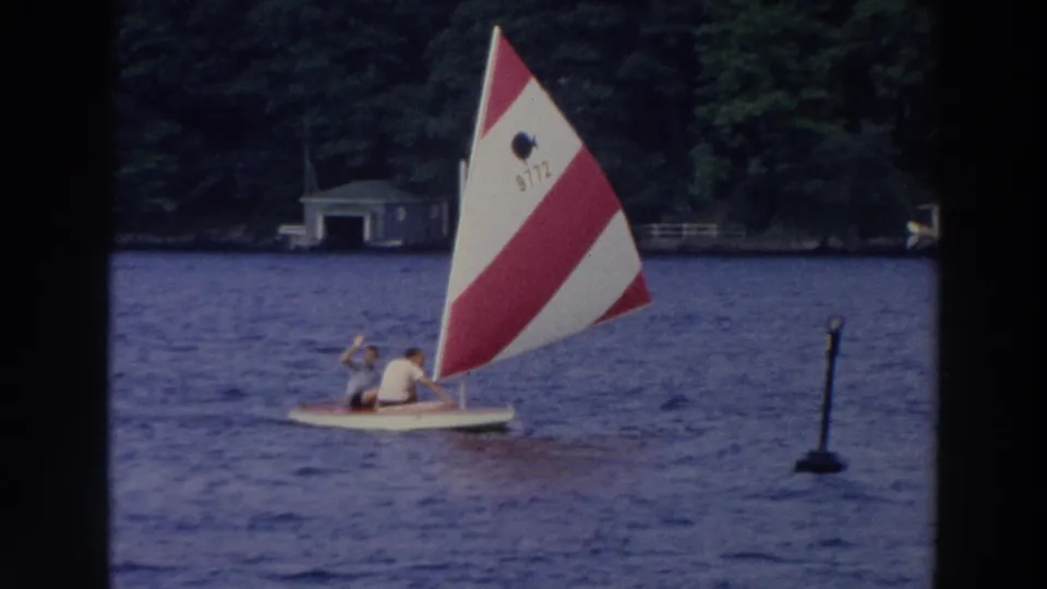 Closer 1966 Wisconsin home-movie view of two people in a small red-and-white striped sailboat near a buoy and wooded shore.