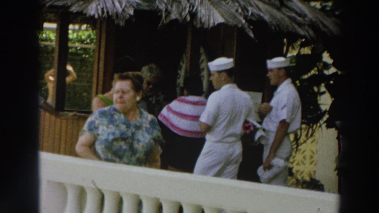 Woman in a blue floral shirt stands by a white railing as sailors in white uniforms walk behind her.