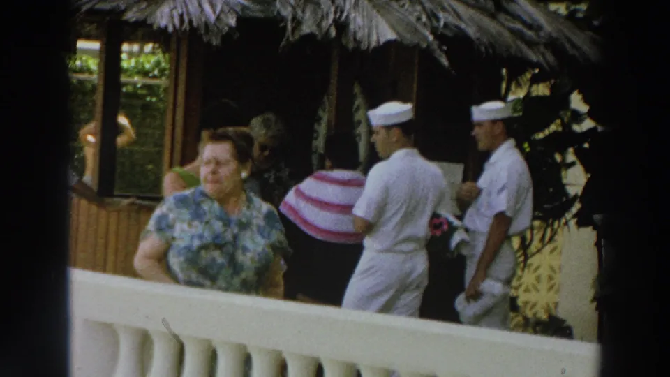 1966 Wisconsin home-movie frame showing a woman near a balcony rail with Navy sailors passing under a thatched roof.