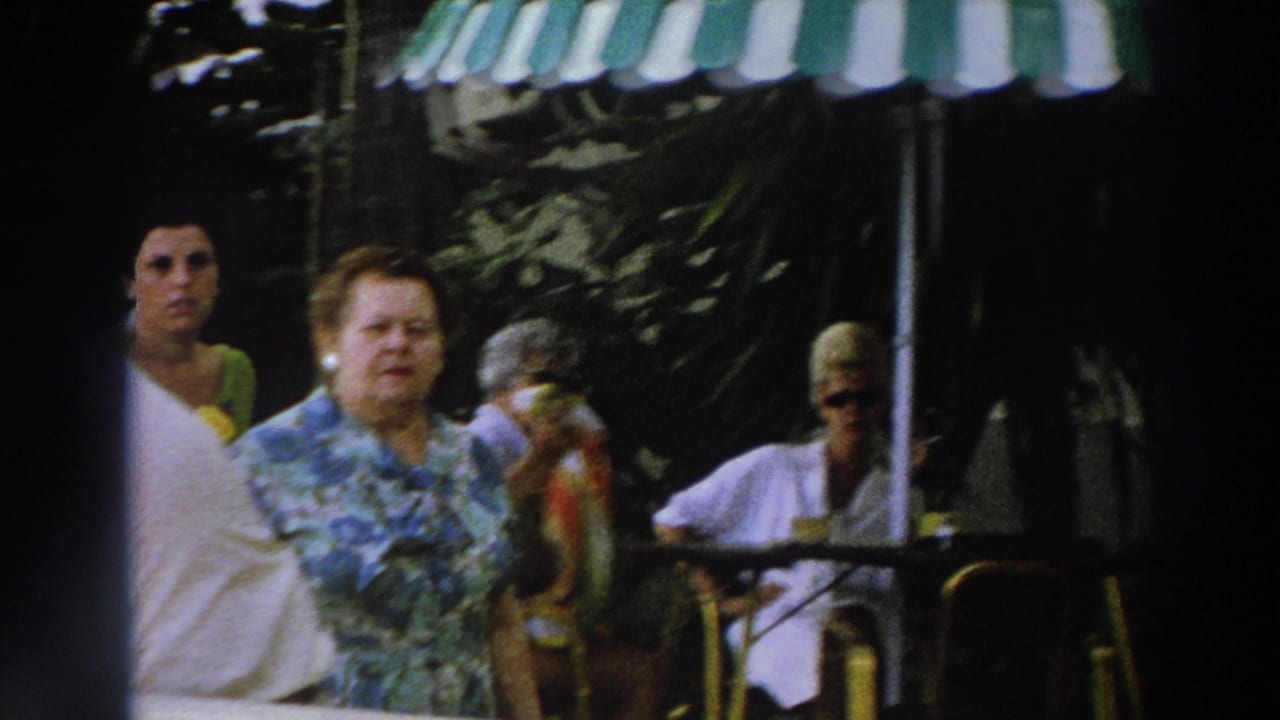 1966 Wisconsin home-movie frame showing two women walking past seated visitors under green-and-white striped awnings.