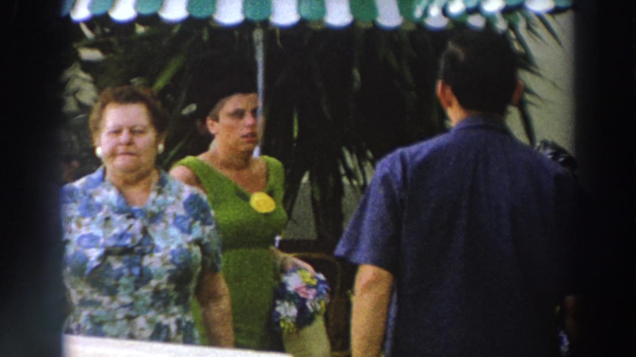 1966 Wisconsin frame with two women in period clothing walking through an outdoor venue near shaded seating.