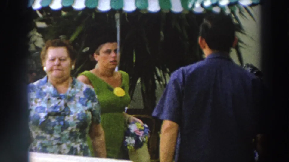 1966 Wisconsin frame with two women in period clothing walking through an outdoor venue near shaded seating.