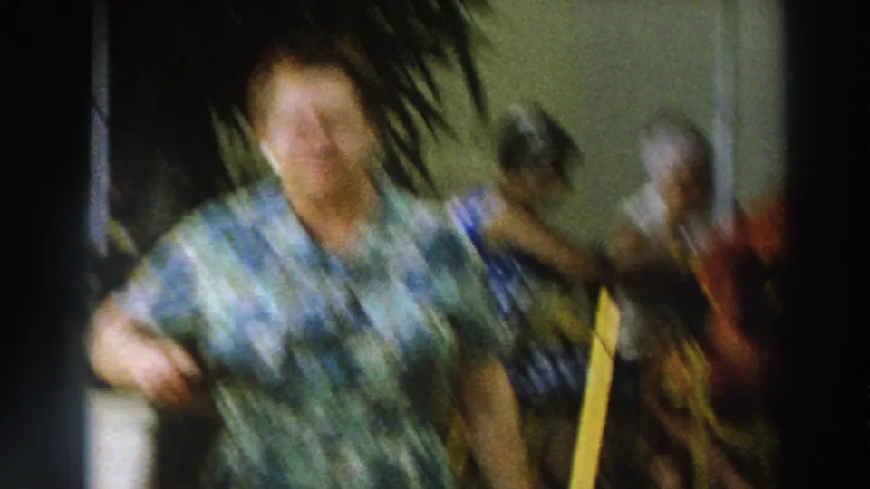 Motion-blurred 1966 Wisconsin frame with a woman moving close to the camera and visitors seated behind her.