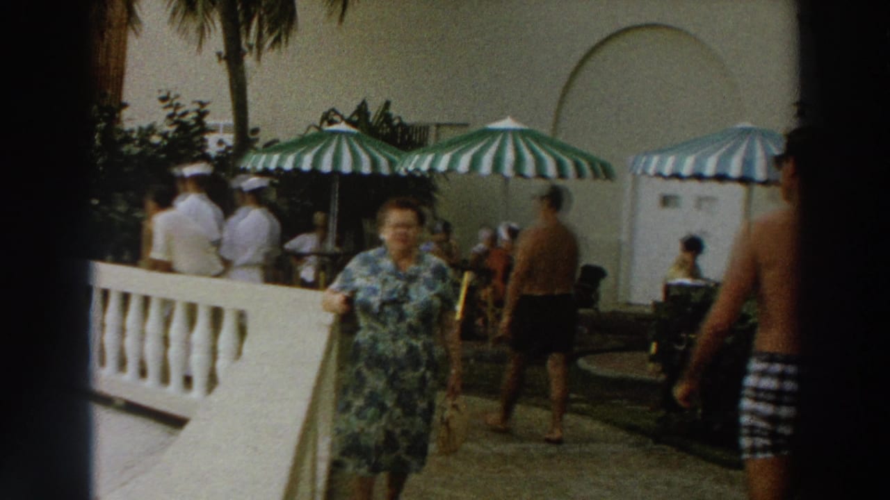 Woman in a floral dress walks toward the camera on a patio with striped umbrellas and guests behind her.