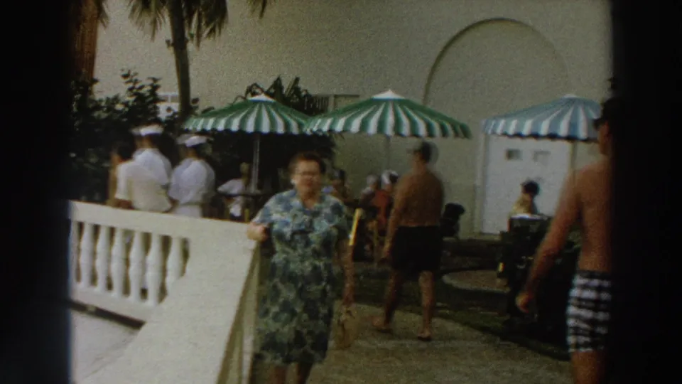 1966 Wisconsin home-movie frame of a woman in a floral dress approaching the camera on a sunlit resort patio with striped umbrellas.