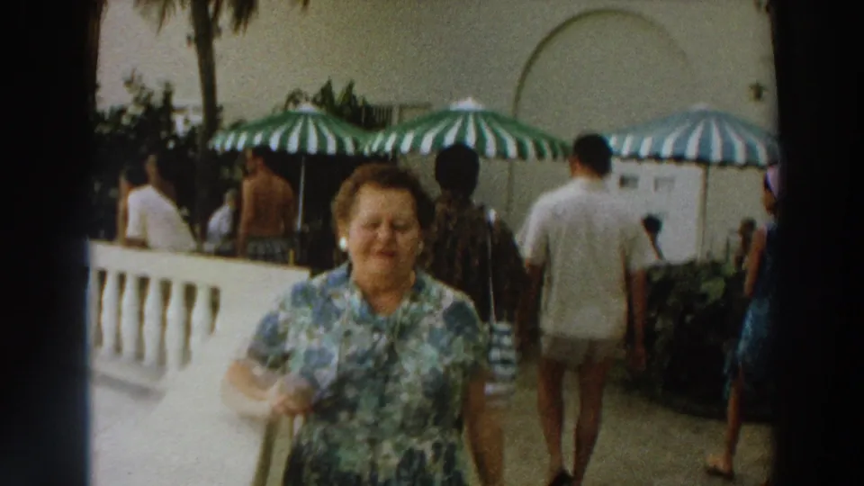 Close 1966 Wisconsin home-movie view of a woman on a resort patio, with adults walking and striped umbrellas in the background.