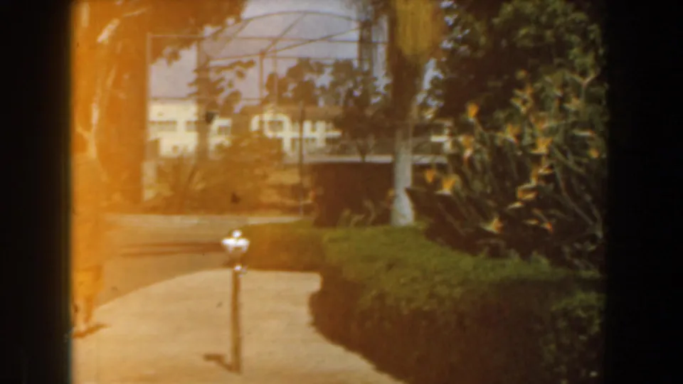 Soft 8mm frame of a Burbank garden path bordered by hedges and shrubs, with white posts and a tennis court structure in the background.