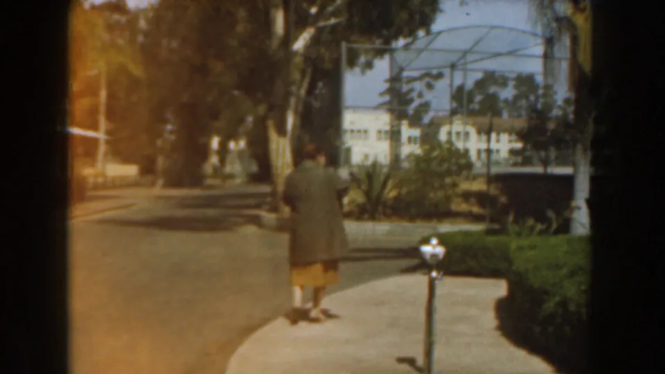 1961 Burbank home-movie frame of a woman walking away on a garden path, with hedges, trees, white posts, and a tennis court nearby.