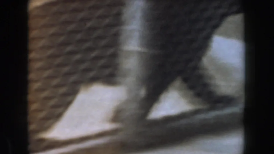 A small dog moves quickly across a light interior floor beside a striped rug or mat in Burbank home-movie footage.