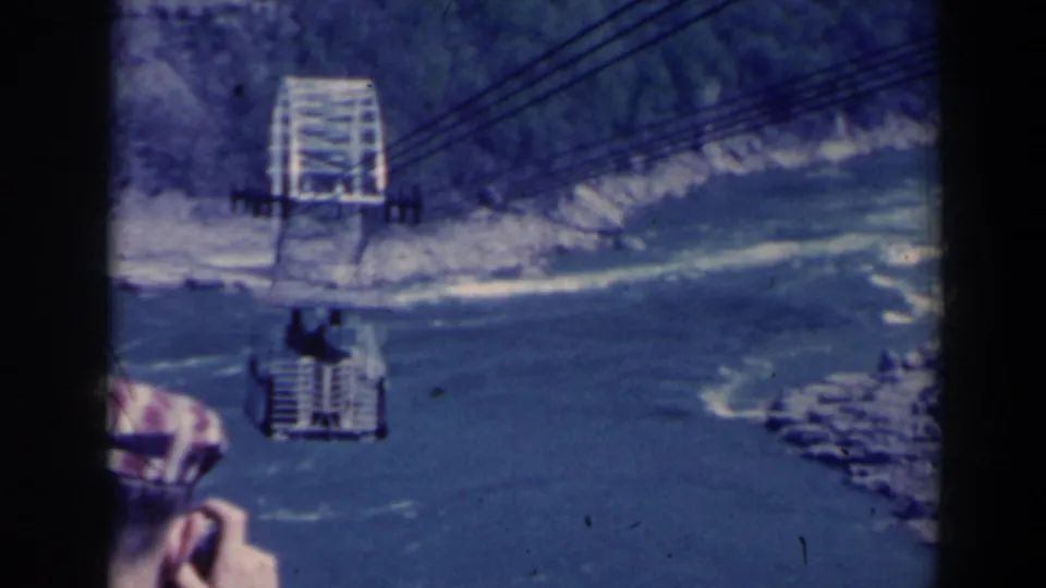 Blue-tinted 8mm frame of a Niagara Gorge cable car over churning water, with terminal structure, cables, rocks, and a partial passenger near the edge.
