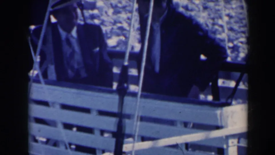 Passengers in an open cable car cabin ride across the Niagara Falls area, with a suited man visible near the center.