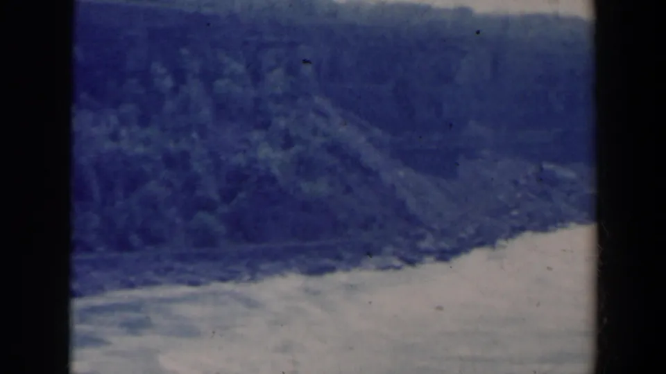 1963 8mm landscape frame showing pale river water below a forested bank, likely near Niagara Falls, Canada, with blue fading and contact wear.