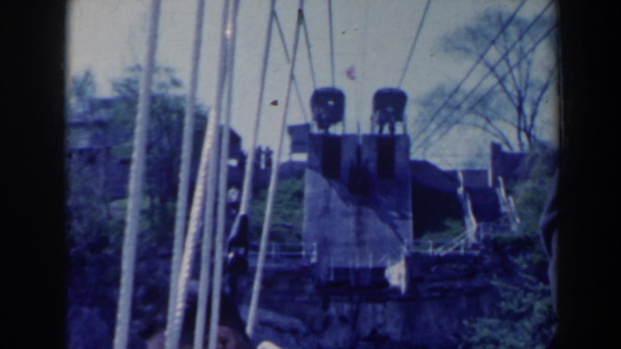Industrial machinery, vertical cables, railings, and shrubs near a bridge-like structure.