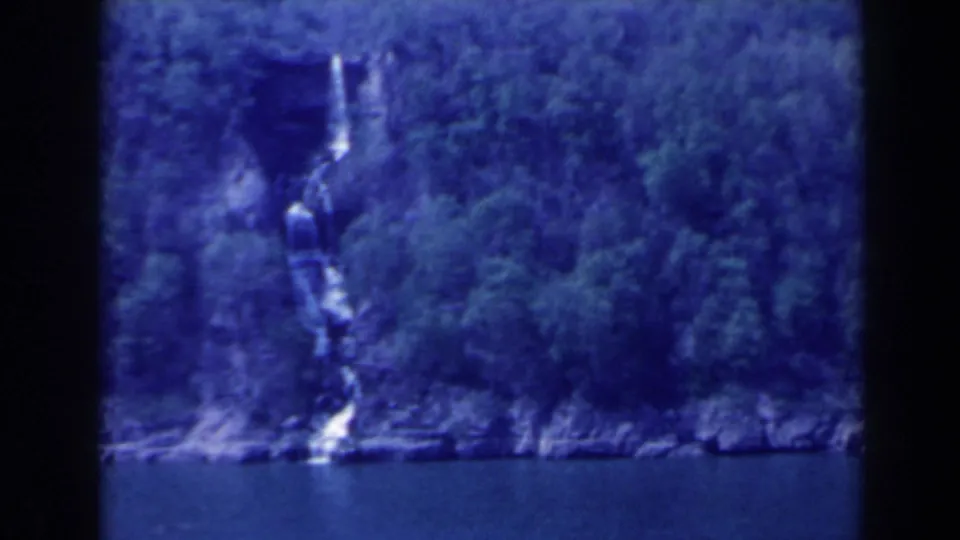 1963 Canada 8mm frame showing a distant waterfall cutting through forested rock above calm water.
