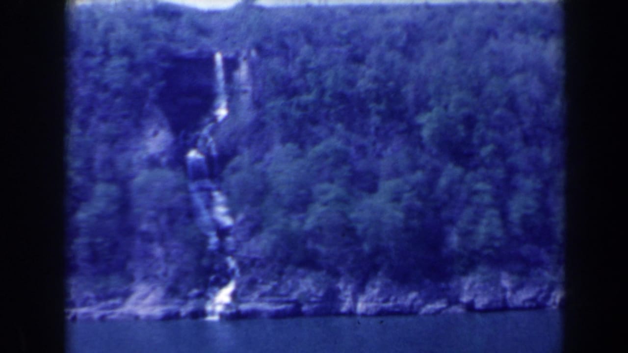 1963 8mm frame of a narrow waterfall on a forested cliff near water in the Niagara Falls, Canada area, with soft focus and blue color cast.