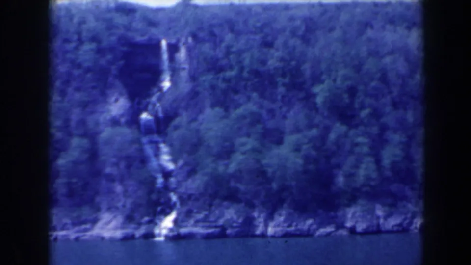 1963 8mm frame of a narrow waterfall on a forested cliff near water in the Niagara Falls, Canada area, with soft focus and blue color cast.