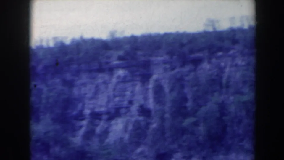 1963 home-movie frame showing a forested cliff face and shoreline near Niagara Falls, Canada, with the waterfall less distinct in the soft scan.