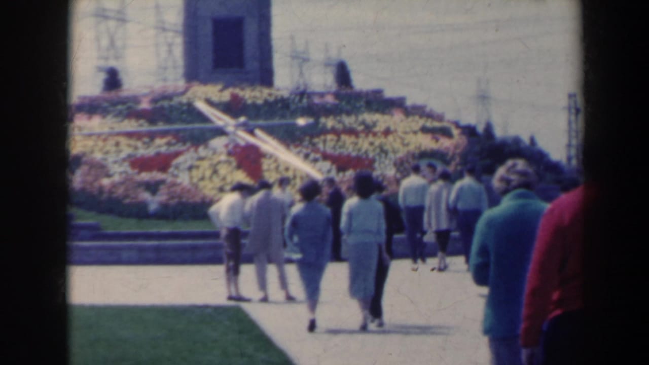 Visitors walk along a paved path toward colorful flower beds and a monument facade.
