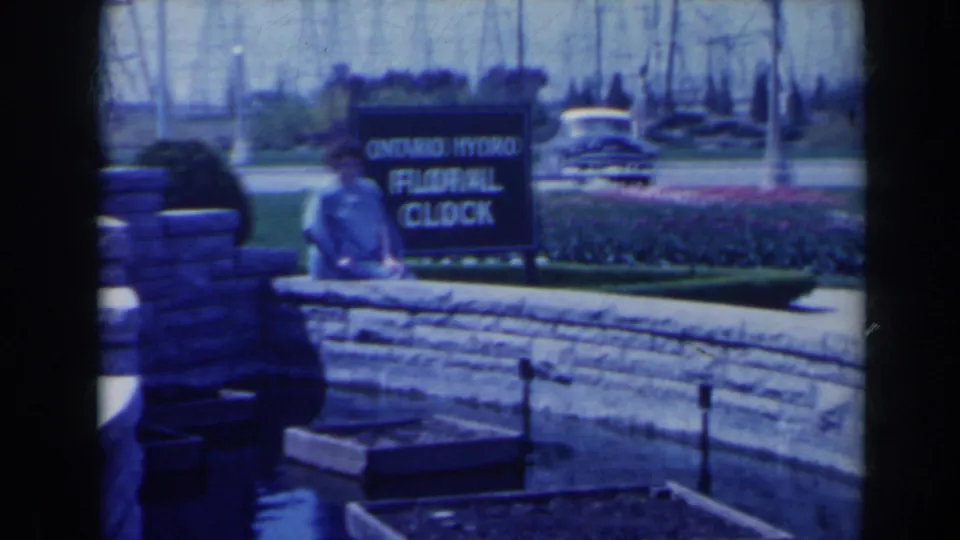 A woman stands by the stone wall and Ontario Hydro Floral Clock sign near tulip beds in Niagara Falls, Canada, 1963.