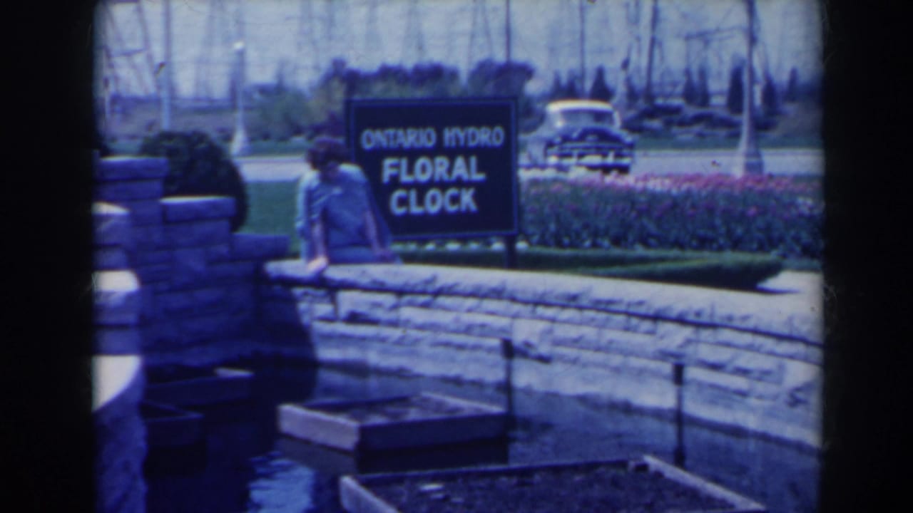 1963 Niagara Falls home-movie frame showing a woman by the Ontario Hydro Floral Clock sign, with flowers, lawn, stonework, and a car in view.