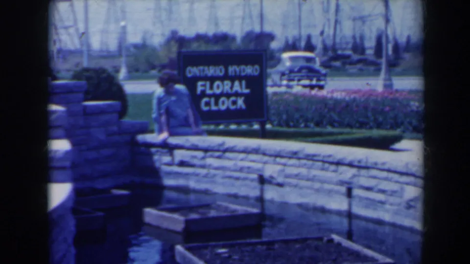 1963 Niagara Falls home-movie frame showing a woman by the Ontario Hydro Floral Clock sign, with flowers, lawn, stonework, and a car in view.