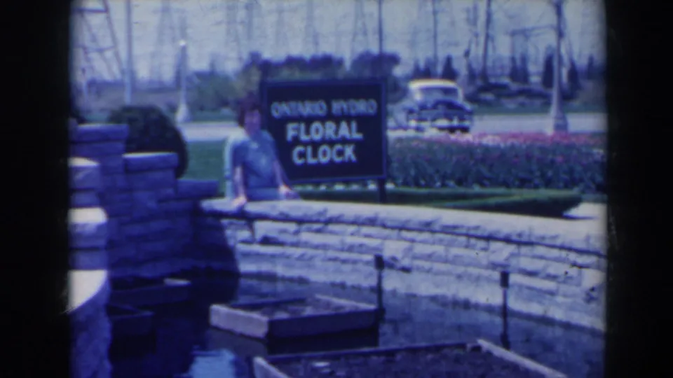 1963 Niagara Falls frame of a woman walking near the Ontario Hydro Floral Clock sign with planted beds, stone wall, and vintage car behind.