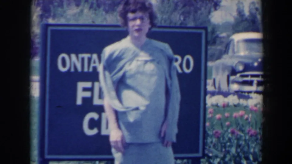 Closer 1963 home-movie view of a woman in a blue dress passing in front of the Ontario Hydro Floral Clock sign at Niagara Falls.