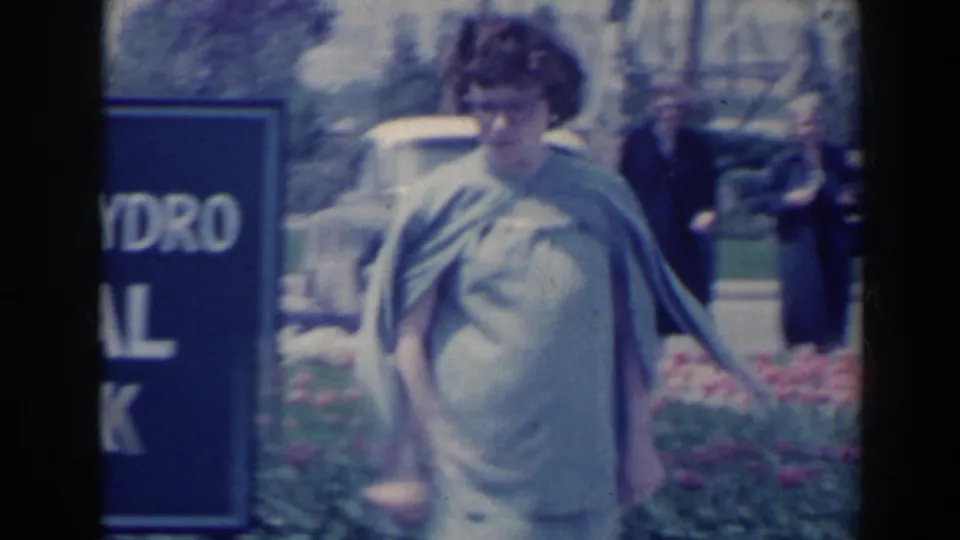 1963 Niagara Falls frame showing the woman walking past the Floral Clock area, with flowers, sign edge, car, and another adult visitor behind.