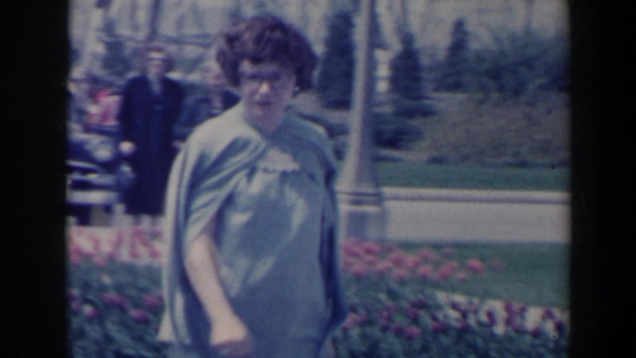Woman in a pale dress and shawl walking past flower beds in a sunny park.