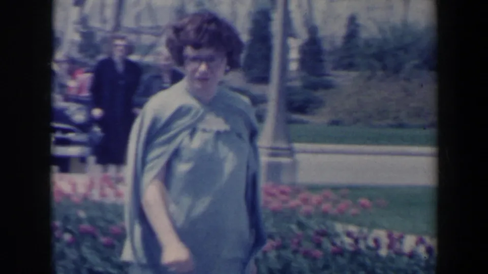 1963 8mm frame of a curly-haired woman in a pale dress and shawl walking in a Niagara Falls, Canada park area.