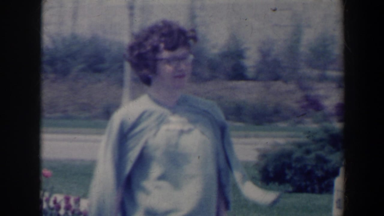 Woman in pale clothing standing near lawns and shrubs with a light shawl moving beside her.