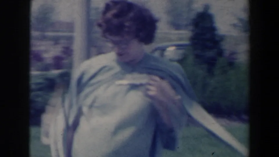 A close, soft 8mm frame shows the woman adjusting her shawl while standing near flower beds and greenery.