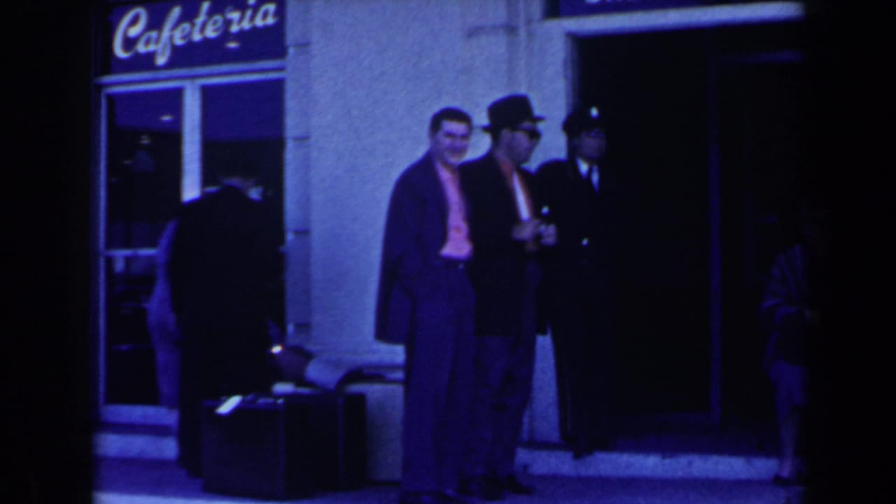 Two suited men stand near luggage outside a hotel entrance as others pass nearby.