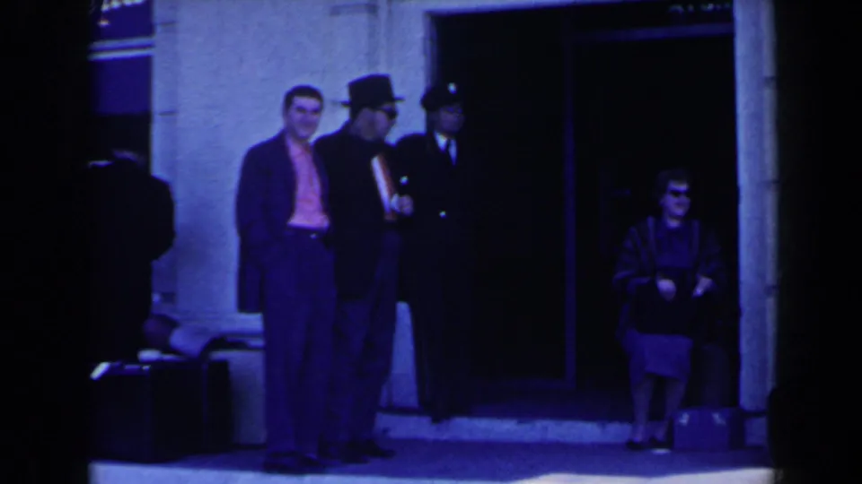 1963 Niagara Falls, Canada home-movie frame showing travelers waiting outside the Sheraton-Brock doorway, including a seated woman with bags.