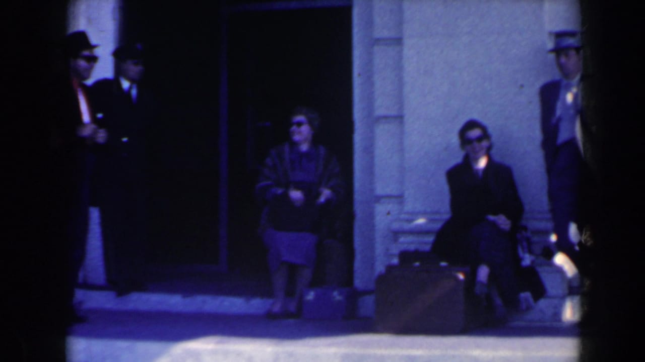 1963 Niagara Falls home-movie frame showing adults in sunglasses and hats waiting near a building entrance with suitcases.