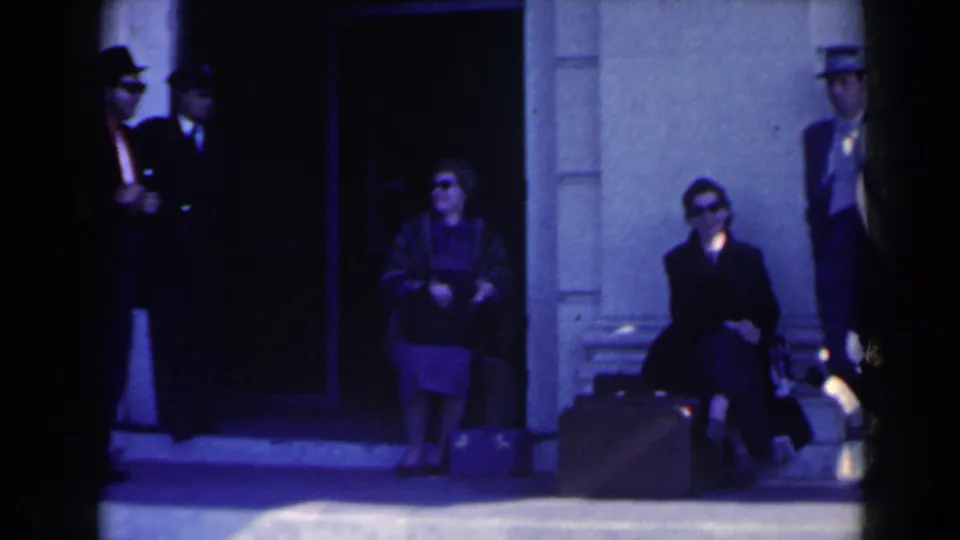 1963 Niagara Falls home-movie frame showing adults in sunglasses and hats waiting near a building entrance with suitcases.