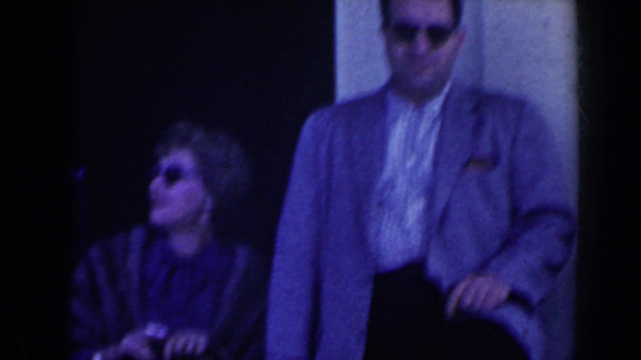 A man in sunglasses stands near a seated woman outside a doorway.