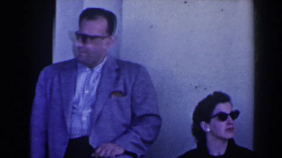 1963 Niagara Falls frame showing a suited man standing beside a seated woman in dark sunglasses against a pale exterior wall.